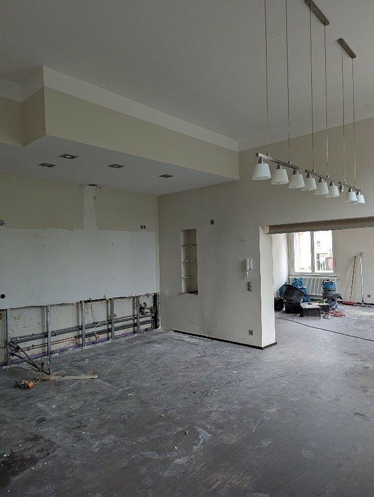 Ein großer Raum während Renovierungsarbeiten. An einer Wand sind die Anschlüsse und Leitungen einer entfernten Küchenzeile freigelegt. Die Decke hat eine abgehängte Konstruktion mit eingelassenen Spots, und mehrere Lampen hängen von der Decke. Der Boden ist staubig und unfertig, Baugeräte stehen verteilt im Raum.
