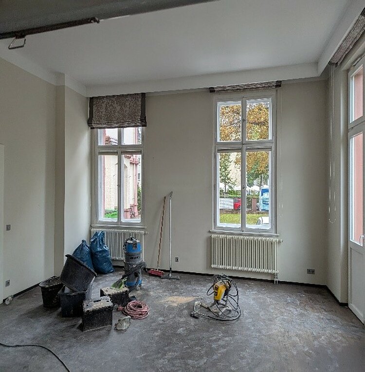 Ein heller Raum im Umbauzustand mit hohen weißen Wänden und großen Fenstern. Auf dem Boden stehen Werkzeuge, Eimer, Müllsäcke und Baumaschinen. Der Boden ist noch unfertig, und die Heizkörper unter den Fenstern sind sichtbar.