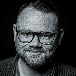 Schwarz-weiß-Porträt eines Mannes mit Brille, kurz geschnittenem Haar und Bart. Er trägt ein gestreiftes Hemd und lächelt leicht in die Kamera vor schwarzem Hintergrund.