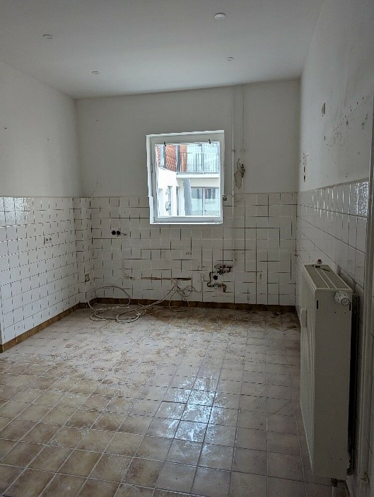 Ein kleiner Raum mit weiß gekachelten Wänden und einem schmutzigen Fliesenboden im Renovierungszustand. Die Anschlüsse einer ehemaligen Küche oder Waschküche sind freigelegt, und Kabel liegen lose am Boden. Unter dem Fenster befindet sich ein Heizkörper, durch das Fenster sind benachbarte Gebäude zu sehen.