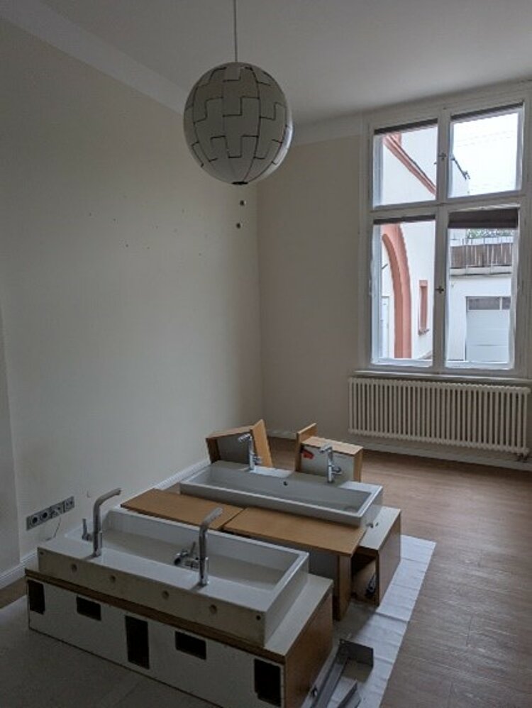 Ein renovierter, heller Raum mit großen Fenstern und einem hängenden, kugelförmigen Lampenschirm. In der Mitte stehen zwei nicht montierte Waschbeckenmodule auf dem Boden bereit zur Installation. Der Raum hat beige Wände und einen hellen Bodenbelag.