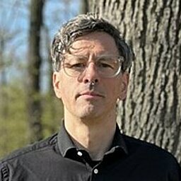 Mann steht in einem Waldgebiet vor einem dicken Baum. Er trägt eine schwarze Brille und ein schwarzes Hemd. Der Hintergrund ist lichtdurchflutet mit kahlen Bäumen.