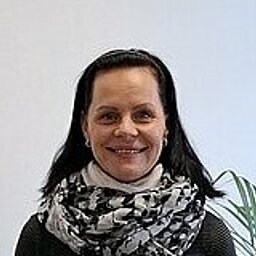 Frau steht lächelnd neben einem Informationsständer mit Broschüren und dem Logo „ESO“. Sie trägt einen schwarzen Pullover, eine helle Hose und einen gemusterten Schal. Im Hintergrund ist eine große Fensterfront mit Ausblick auf ein Bürogebäude und eine Topfpflanze.