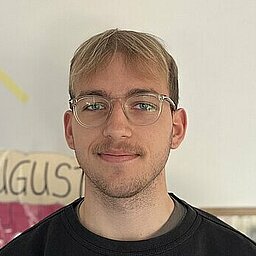 Ein junger Mann mit Brille, blondem Haar und kurzem Bart steht vor einer hellen Wand mit bunten Dekorationen, darunter die Monatsnamen „Juli“ und „August“. Er trägt einen schwarzen Nike-Pullover und blickt freundlich in die Kamera. Im Hintergrund ist ein Teil eines Klassen- oder Gruppenraums zu erkennen.