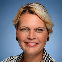 Porträt einer lächelnden Frau mit blondem Haar vor blauem Hintergrund. Sie trägt ein schwarz-weiß gestreiftes Jackett über einem weißen Oberteil und eine lange Kette.